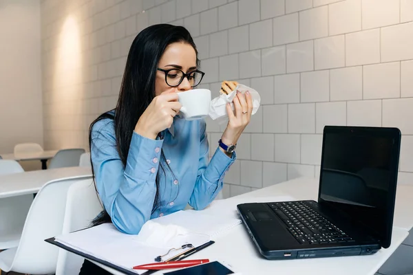 Chica ocupada estudiante en vasos de trabajo en una cafetería con un ordenador portátil, un desayuno buffet con un sándwich y café — Foto de Stock