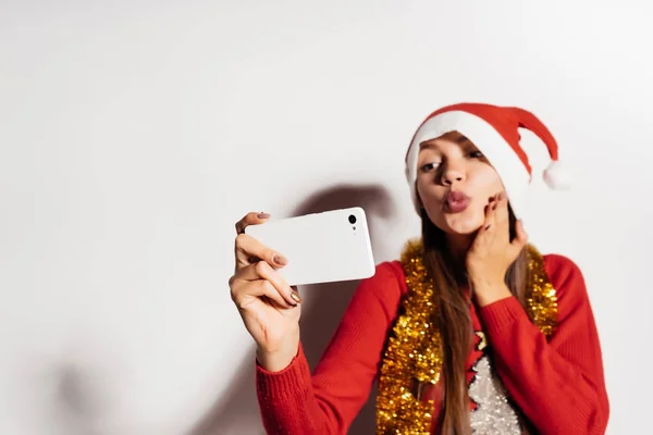 Ein junges Mädchen macht ein Selfie, in festlicher Kleidung und einer Mütze wie der Weihnachtsmann, und wartet auf ein neues Jahr und Weihnachten — Stockfoto