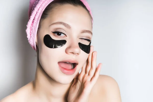 Забавная молодая девушка смотрит за кожей на лице, под глазами увлажняющие пятна, на голове розовое полотенце — стоковое фото