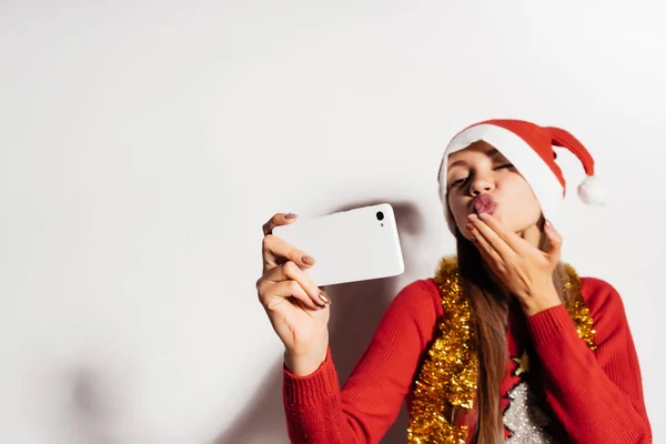 A sweet girl in a festive hat waiting for a new year and Christmas, makes selfie, sends an air kiss to Santa Claus — Stock Photo, Image