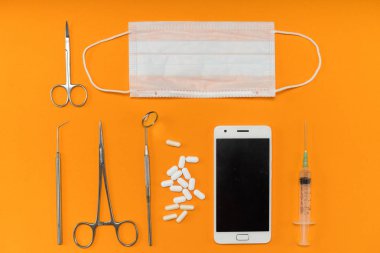 Turuncu yüzeyde bir akıllı telefon, bir şırınga, hapları, diş aletleri ve yüz maskesi olduğunu