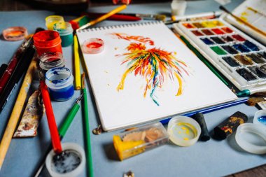 mavi bir masada sanat malzemeleri, kağıt, suluboya ve guaş, fırçalar ve kalem çizim vardır