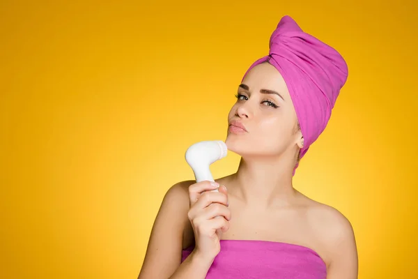 Привлекательная девушка с розовым полотенцем на голове заботится о ее лице, очищает его электрической щеткой — стоковое фото