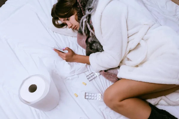 Gadis muda yang sedih sakit, berbaring di tempat tidur, di samping pil berbohong, sakit tenggorokan, terus smartphone — Stok Foto