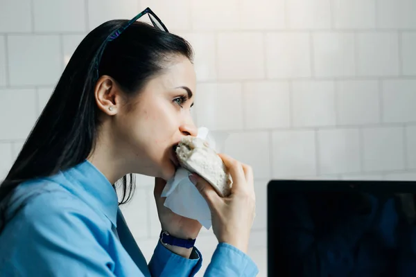Divertida mujer de negocios ocupada en una camisa azul trabajando en un café, comiendo un sándwich — Foto de Stock