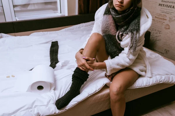 Грустная молодая девушка заболела, сидит на кровати в белом махровом халате, теплый шарф на шее — стоковое фото