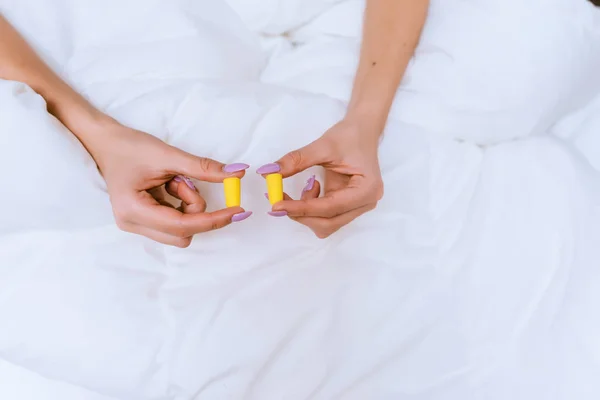 Menina senta-se na cama sob um cobertor branco, mantém tampões de ouvido amarelos contra o ruído — Fotografia de Stock