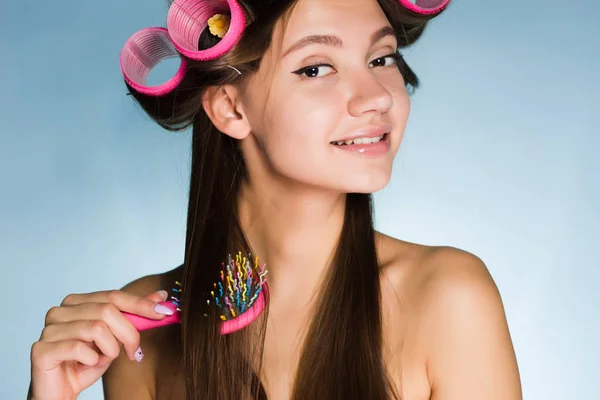 A smiling young girl makes a fashionable hairstyle, on her head a big pink curler, combs her hair — Stock Photo, Image