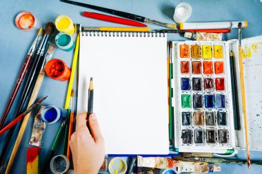 mavi bir masada sanat malzemeleri, kağıt, suluboya ve guaş, fırçalar ve kalem çizim vardır