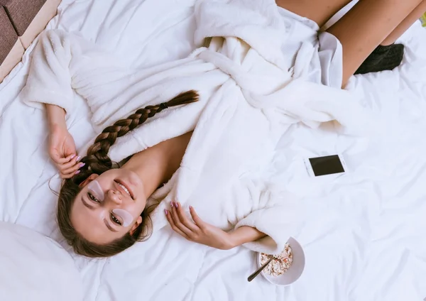 Happy νεαρό κορίτσι βρίσκεται σε ένα κρεβάτι σε μια ρόμπα terry λευκό, απολαμβάνει το Σαββατοκύριακο, κάτω από τα μάτια των patches σιλικόνης, δίπλα σε ένα πιάτο με ένα χρήσιμο Πλιγούρι βρώμης κουάκερ — Φωτογραφία Αρχείου