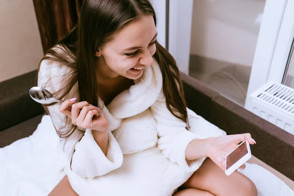 Seorang gadis muda tertawa dalam gaun terry putih melihat ke dalam smartphone-nya — Stok Foto