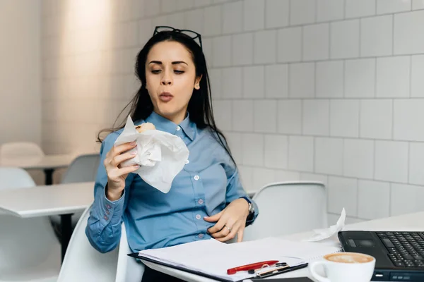 Молодая девушка-фрилансер завтракает в кафе с бутербродом и кофе, работает на ноутбуке — стоковое фото