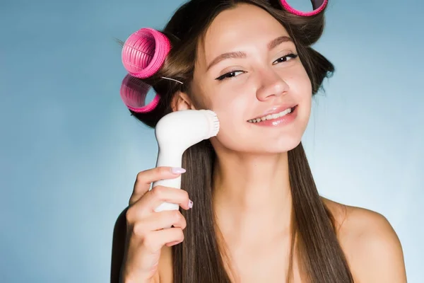 Μια ευτυχισμένη κοπέλα φροντίζει τον εαυτό της, στο κεφάλι το ψαλίδι για μπούκλες του, καθαρίζει το δέρμα στο πρόσωπό της με μια ηλεκτρική βούρτσα — Φωτογραφία Αρχείου