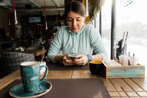 La chica está sentada en un café en una mesa, sosteniendo un teléfono inteligente en sus manos —  Fotos de Stock