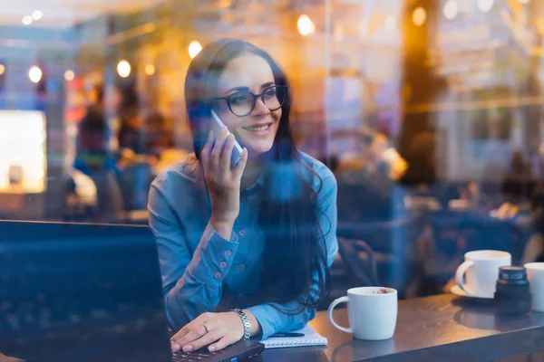 Boldog, sikeres üzletasszony szemüveg ül egy kávézóban, beszél a telefonon — Stock Fotó