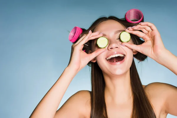 Legrační mladá dívka chce vypadat dobře, na hlavu růžový kulma, udržuje kousky okurky pro hydrataci pokožky — Stock fotografie