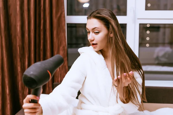 Lovely girl in a white terry robe dries her hair with a hairdryer — Stock Photo, Image