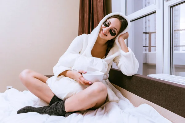 Menina feliz em um vestido de vestir branco terry senta-se na cama, sob os olhos de manchas de silicone preto, tomando café da manhã na cama — Fotografia de Stock