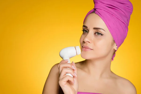 Νεαρό κορίτσι με μια ροζ πετσέτα στο κεφάλι της καθαρίζει το δέρμα στο πρόσωπό της με μια ηλεκτρική βούρτσα — Φωτογραφία Αρχείου