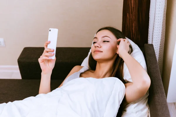 Seorang gadis muda baru-baru ini bangun, berbaring di bawah selimut dan melihat ke dalam smartphone nya — Stok Foto