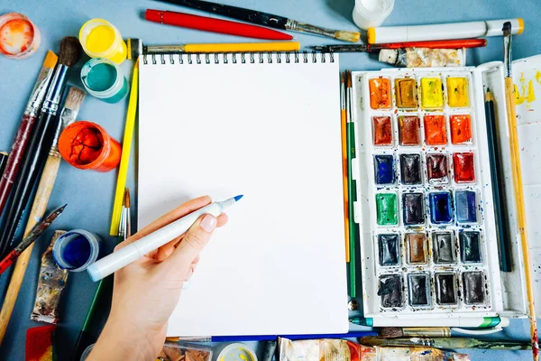 mavi bir masada sanatsal malzemeleri, kağıt, suluboya, guaş, birçok fırçaları vardır