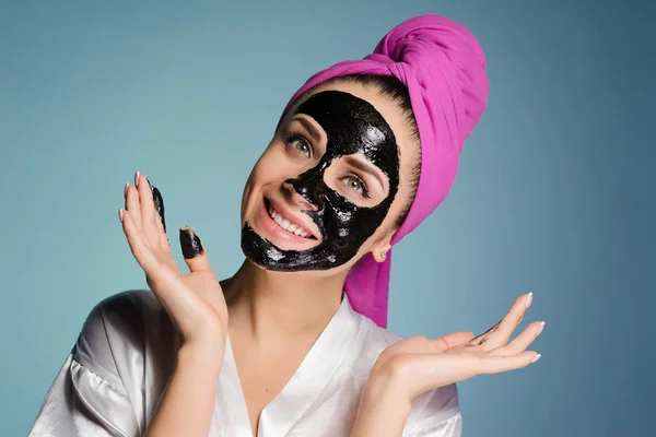 Menina sorridente feliz com uma toalha rosa na cabeça, em seu rosto uma máscara de limpeza preta — Fotografia de Stock