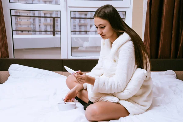 어린 소녀 최근 일어, 백색 테리 드레싱 가운, 침대에 그녀의 스마트폰으로 본다 — 스톡 사진