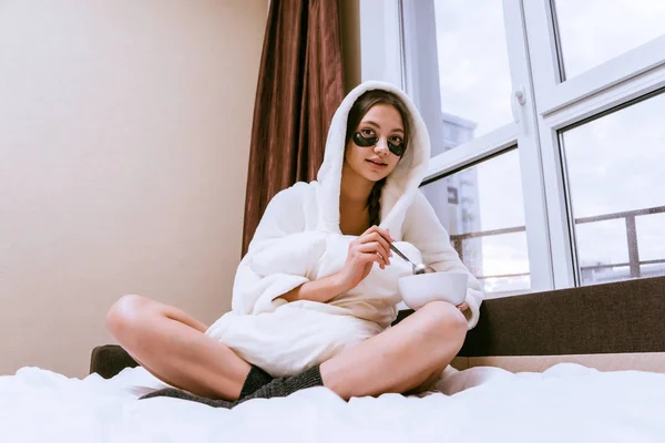 Uma jovem menina em um vestido de vestir branco terry está tomando café da manhã na cama, sob os olhos de manchas de silicone preto — Fotografia de Stock