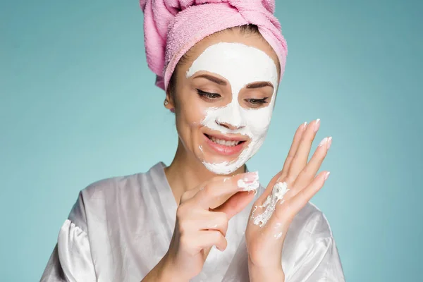 Ένα νεαρό κορίτσι με μια ροζ πετσέτα στο κεφάλι της είναι η εφαρμογή μια λευκή θρεπτική μάσκα στο πρόσωπό της — Φωτογραφία Αρχείου