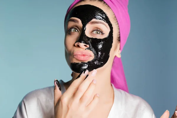 Menina bonita com uma toalha rosa na cabeça cuida da pele, aplica uma máscara de limpeza preta em seu rosto — Fotografia de Stock