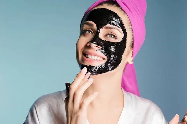 Sorridente ragazza con un asciugamano rosa sulla testa, vuole essere bella, mette una maschera detergente nera sul suo viso — Foto Stock