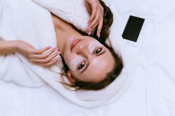 Jovem atraente encontra-se na cama, sob os olhos de manchas de silicone branco, em seguida, é um smartphone — Fotografia de Stock