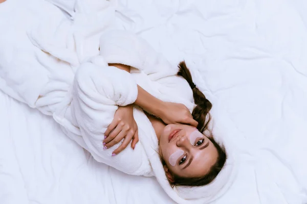 Uma jovem jaz na cama, levantou-se cedo, em um vestido de vestir branco terry, sob os olhos remendos de silicone hidratante — Fotografia de Stock