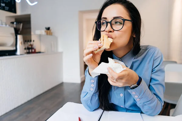 Занятая девушка в очках завтракает в кафе с бутербродом и кофе — стоковое фото