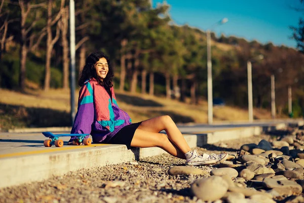 Feliz chica con estilo en una chaqueta púrpura descansa, se sienta al lado de un monopatín — Foto de Stock