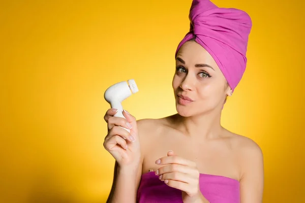 Uma jovem engraçada quer ser bonita, limpa a pele em seu rosto com uma escova elétrica, em um fundo amarelo — Fotografia de Stock