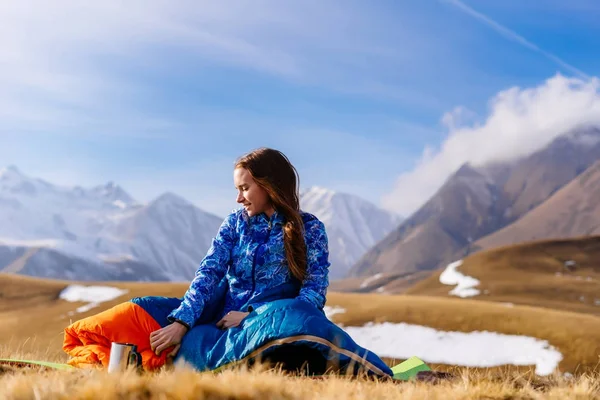 Счастливая молодая девушка в голубой куртке сидит на фоне Кавказских гор, наслаждается природой и теплой погодой — стоковое фото