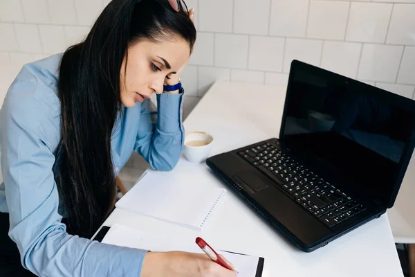 Κουρασμένος απασχολημένος μελαχρινή κοπέλα εργασία σε καφενείο στον φορητό υπολογιστή, γράφοντας κάτι σε χαρτί — Φωτογραφία Αρχείου