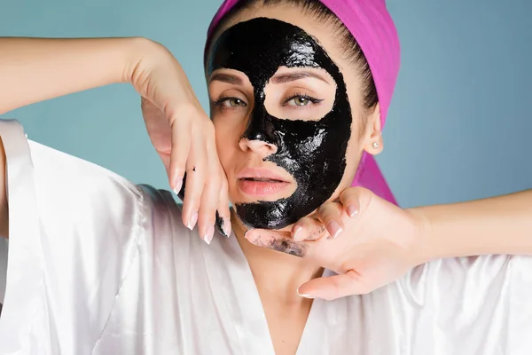 Uma jovem atraente colocar uma máscara de limpeza preta em seu rosto, em sua cabeça uma toalha rosa — Fotografia de Stock