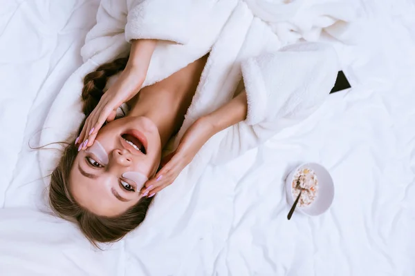 Dziewczyna obudził się wcześnie, leżąc na łóżku w białym płaszczu, pod jej oczy silikonowe plastry przeciw niebieskie kółka — Zdjęcie stockowe