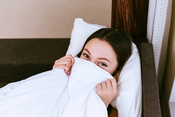 Lustiges junges Mädchen liegt im Bett, bedeckt das halbe Gesicht mit weißer Decke — Stockfoto