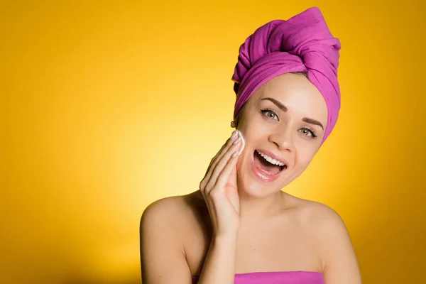 Glad ung flicka med rosa handduk på huvudet rengör huden i ansiktet med en bomull-skiva — Stockfoto