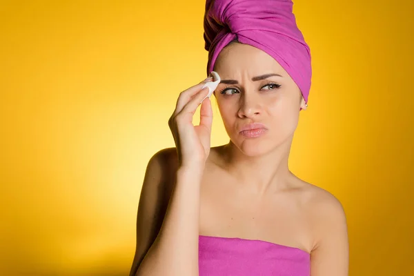 Menina envergonhada descontente com uma toalha rosa na cabeça limpa a pele em seu rosto com um disco de algodão — Fotografia de Stock