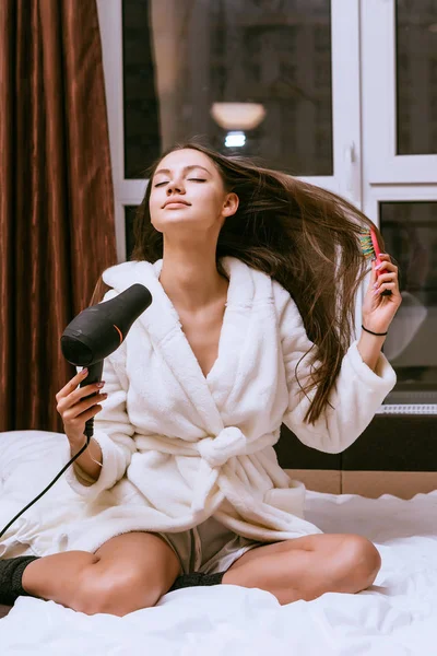Een mooi jong meisje in een witte terry gewaad droogt haar lange haren met een haardroger — Stockfoto
