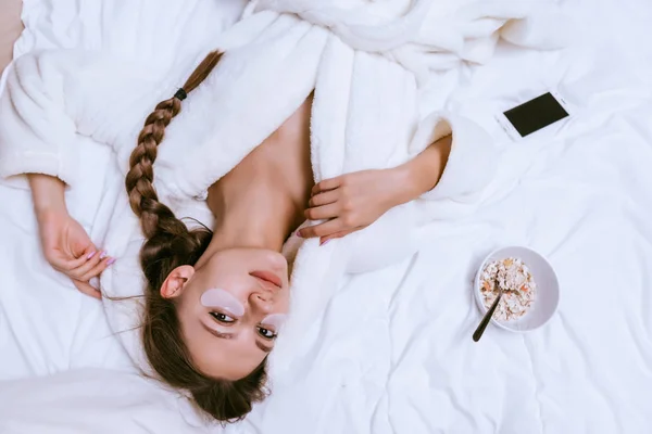 Όμορφη νεαρή κοπέλα σε έναν λευκό terry ρόμπα βρίσκεται στο κρεβάτι, κάτω από τα μάτια των patches άσπρη σιλικόνη, δίπλα σε ένα πιάτο με αλεύρι βρώμης κουάκερ — Φωτογραφία Αρχείου