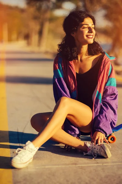 Felice ragazza elegante in una giacca viola siede sul suo skateboard nei raggi del sole della sera — Foto Stock