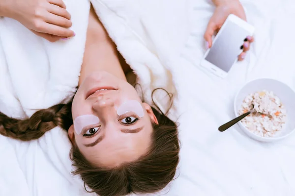 Mooi jong meisje ligt op een bed, onder de ogen van witte siliconen patches, naast een plaat met een nuttige havermoutpap — Stockfoto
