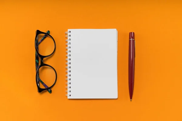 Na oranžové pozadí leží bílý notebook, červené pero a černém obroučkami — Stock fotografie