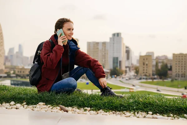 Elegante ragazza sorridente seduta su una radura verde in un parco e parlare al telefono — Foto Stock