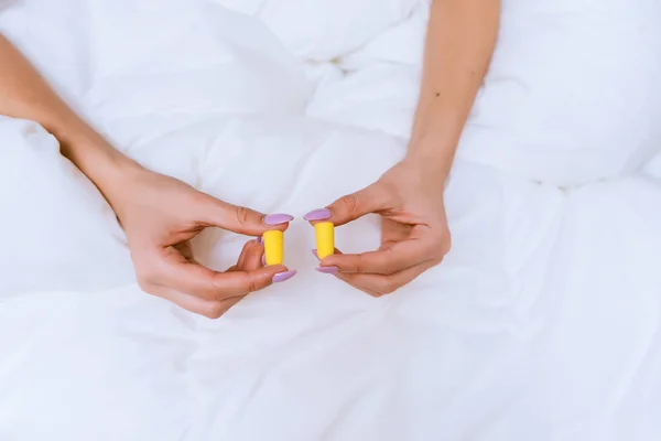 Meisje zit in bed onder een witte deken, gele oordoppen houden tegen straatrumoer — Stockfoto